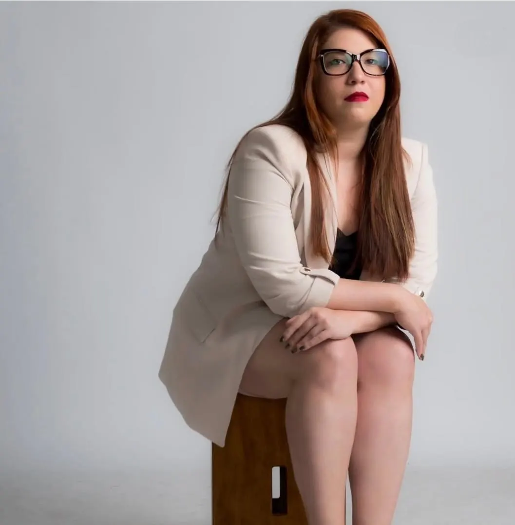 Michelle Santiago, de óculos e blazer claro, sentada num banco de madeira em estúdio claro