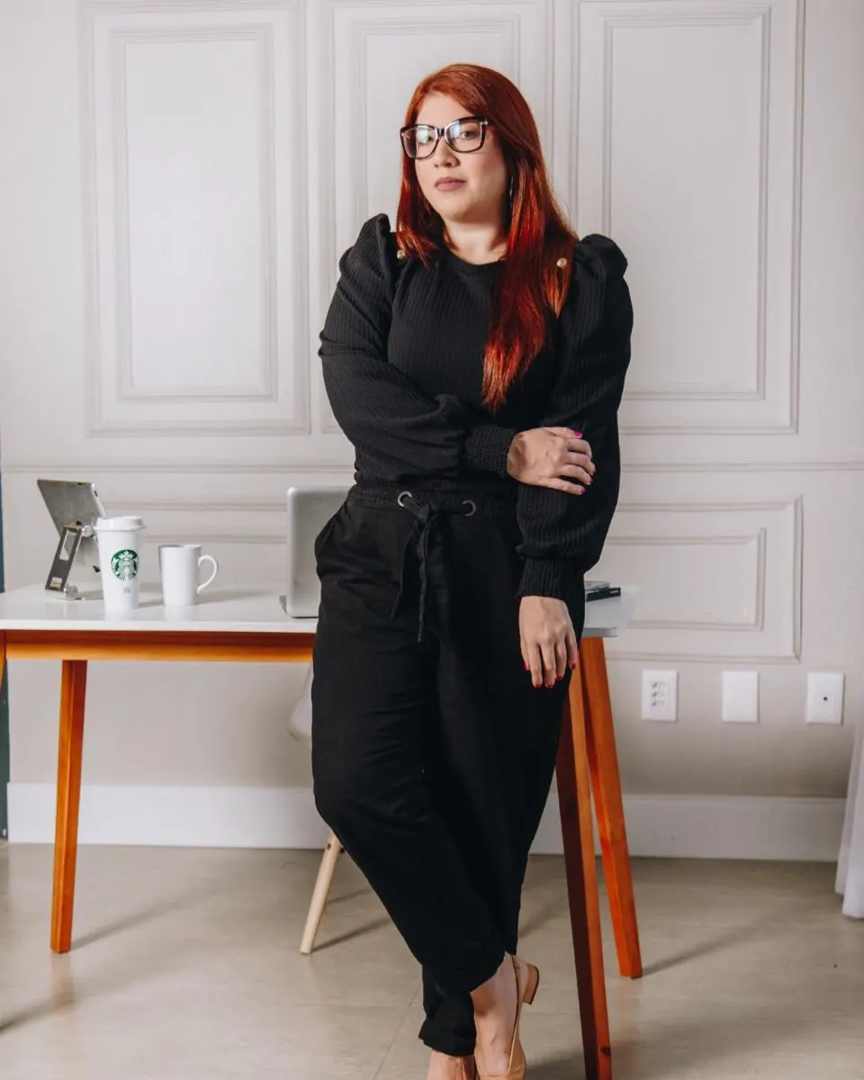 Michelle Santiago em pé, de roupa preta, encostada numa mesa de trabalho
