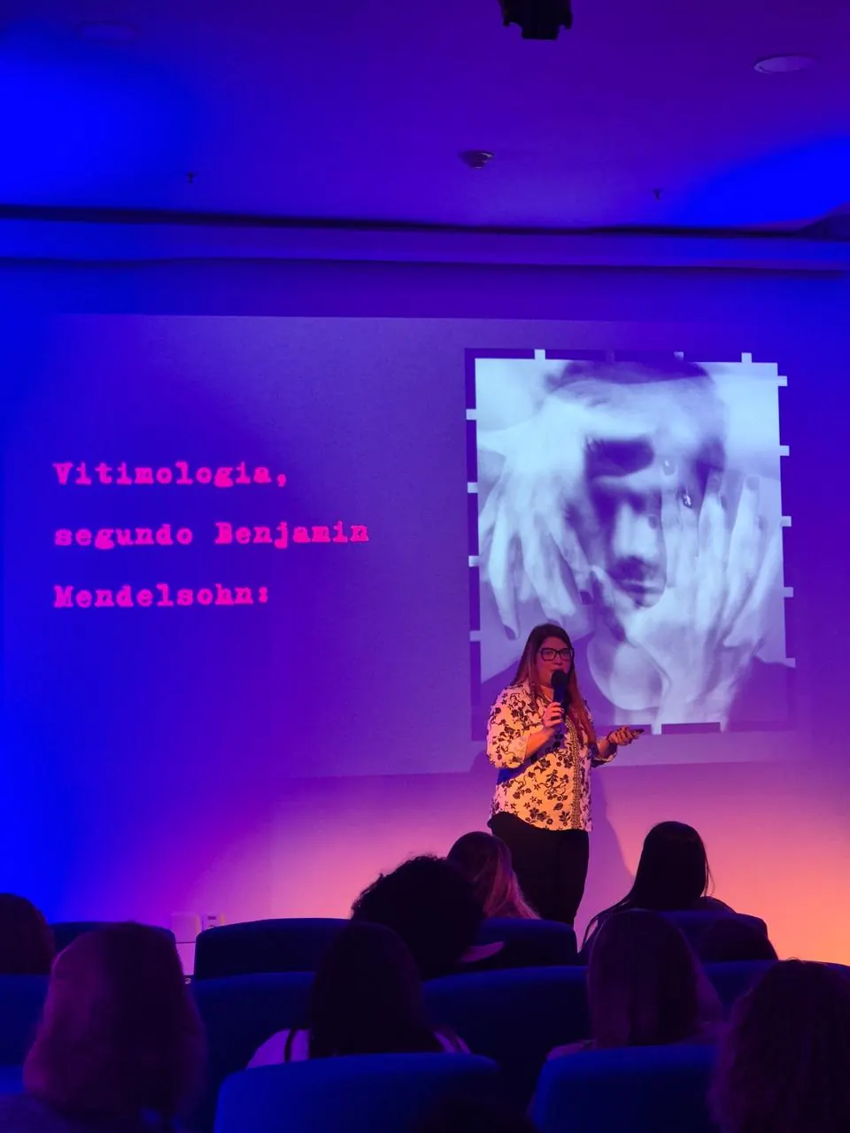 Michelle Santiago no palco, sob luz roxa, apresentando uma palestra sobre vitimologia